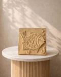 A 6″ x 6″ Terracotta Fish Wall Tile featuring an engraved fish and bubbles sits on a round marble pedestal, set against a neutral textured wall with soft shadow patterns.