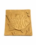 A 6″ x 6″ Terracotta Fish Wall Tile featuring an engraved fish and bubbles sits on a round marble pedestal, set against a neutral textured wall with soft shadow patterns.