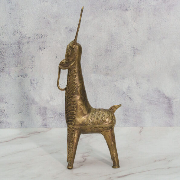 A brass figurine of a stylized animal, possibly a horse or unicorn, with an elongated body, upright horn, and decorative patterns, stands on a light marble surface against a textured pale background.