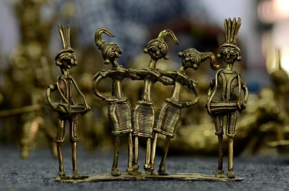 Four handcrafted brass figurines with elongated limbs and expressive faces stand together, each with a unique hairstyle and attire, displayed on a surface with a blurred background.