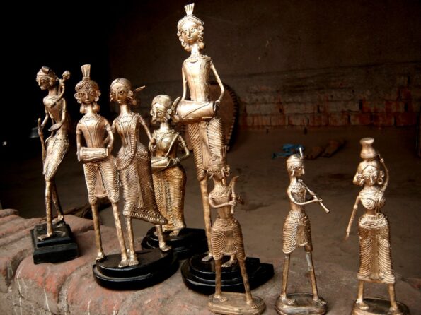 A group of traditional brass figurines, crafted in an indigenous style, stand on a brick surface against a dark background. The figures represent musicians and dancers, each holding various instruments or posing gracefully.