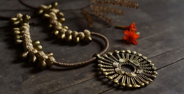 A rustic, handcrafted brass necklace with intricate detailing and a large circular pendant lies on a wooden surface beside dried leaves and an orange flower.