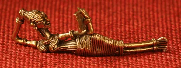 A small brass figurine of a person lying down and reading a book, crafted in a traditional tribal art style, placed on a textured red fabric background.